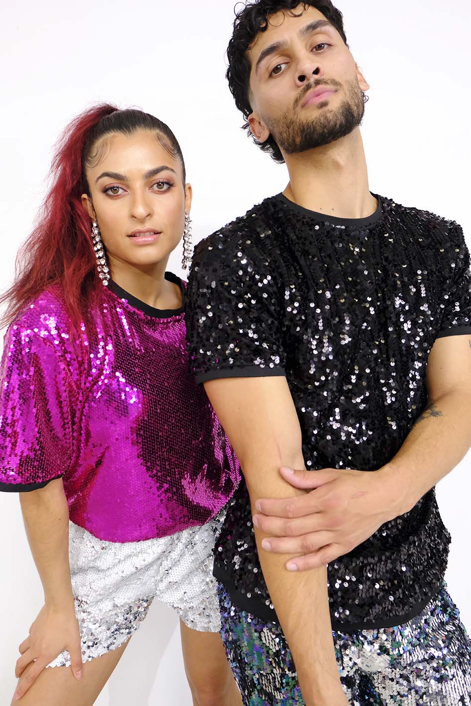 man wearing black sequin t-shirt and purple blue sequin shorts as part of a rave outfit for men. Woman wearing pink sequin t-shirt and white silver sequin shorts as part of a festival outfit for women
