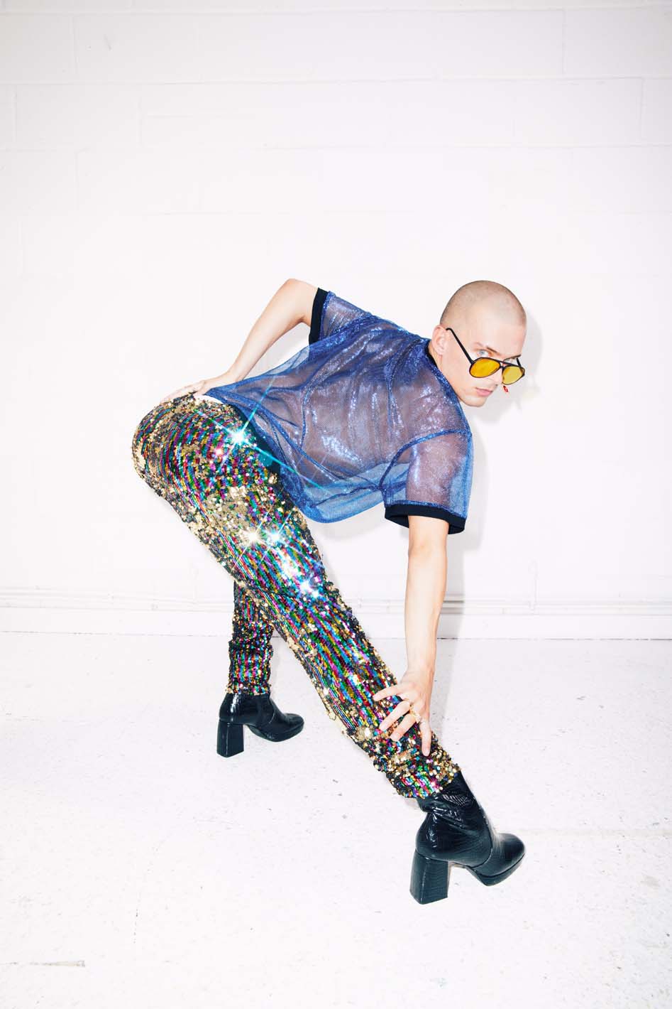 man wearing rainbow sequin pants, blue sheer t-shirt, heels and yellow glasses for gay pride event